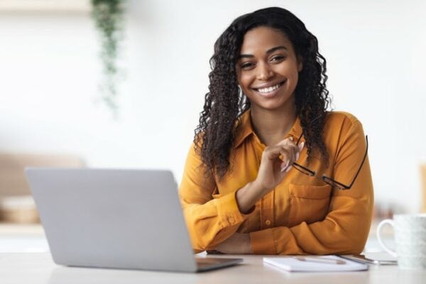smiling teacher wearing yellow blouse and holding eyeglasses while sitting at table with laptop