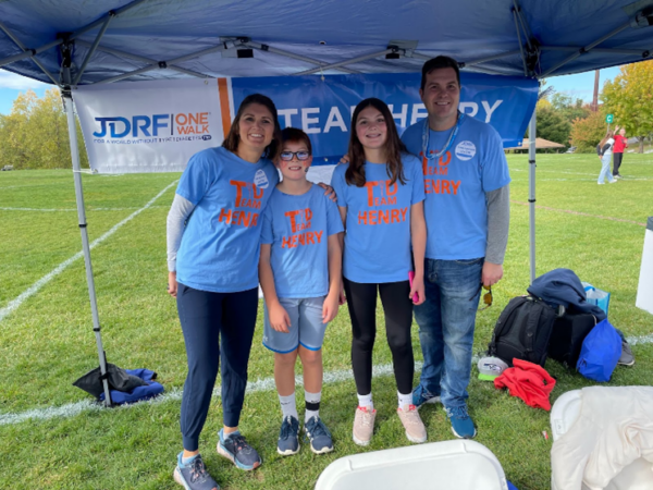 2 adults 2 children in light blue Team Henry tshirts standing outside by Team Henry banner under a canopy tent