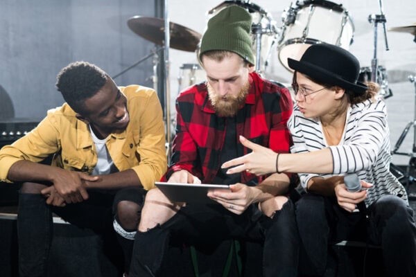 Portrait of three contemporary musicians using digital tablet while discussing songwriting in rehearsal studio