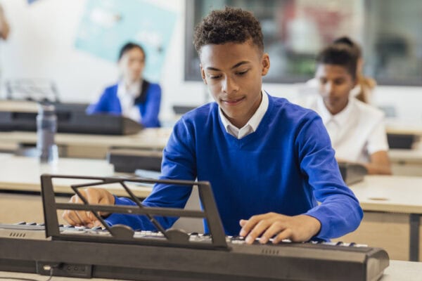 A shot of teenage male music student in a classroom learning how to play the keyboard at a school. The students are all wearing school uniforms.