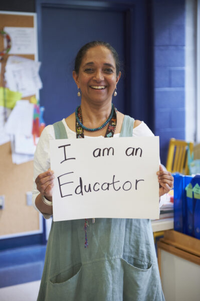 older smiling teacher holding sign reading I am an Educator