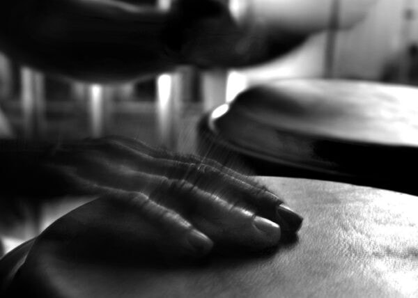 A grayscale image of a hand rhythmically playing a drum, with motion blur emphasizing dynamic movement. The scene conveys a sense of energy and focus.