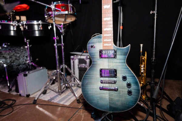A blue electric guitar stands on a stage surrounded by musical instruments, including drums and a trumpet, set against a backdrop of a dark curtain.