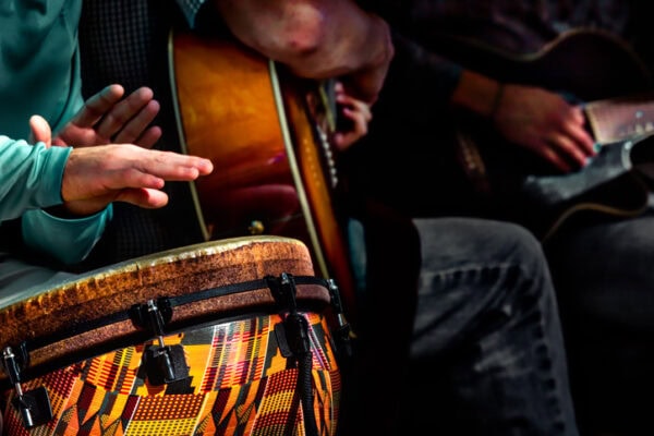Close-up of hands playing drums and guitar. The drum has a colorful, patterned surface. The setting appears energetic and focused on music-making.