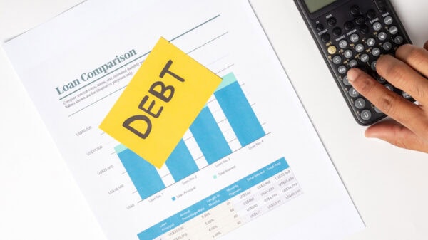 A person is using a calculator to review figures related to debt on a desk. A paper with loan comparison charts and data is nearby, showing various loans and their terms. A bright yellow sticky note is placed on the document, clearly displaying the word DEBT. 
