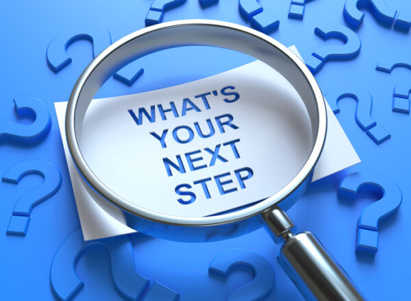 Magnifying glass over note reading Whats Your Next Step? on blue background with question marks