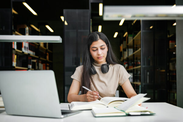 A young woman works diligently with open books and a laptop in a quiet library environment. Her headphones rest around her neck, highlighting a focused and productive study session.