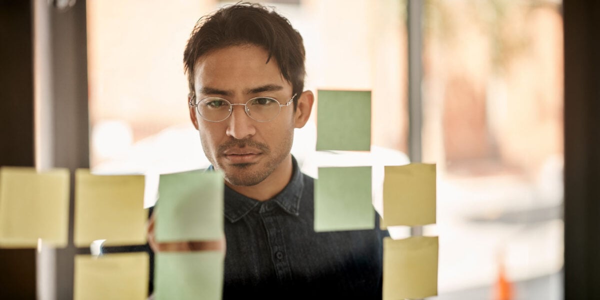 a young man wearing glasses is intently looking at sticky notes he has on a glass wall that he is using for planning