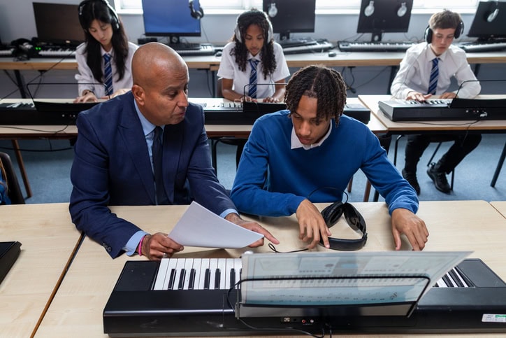 A male teacher in a suit sits next to a teenage schoolboy, guiding him through a sheet music lesson on an electronic keyboard. Other students practice in the background of a secondary school classroom.