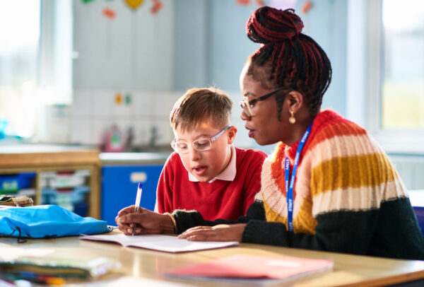Female educational support professional assisting student with disabilities, writing in his book as he watches and concentrates