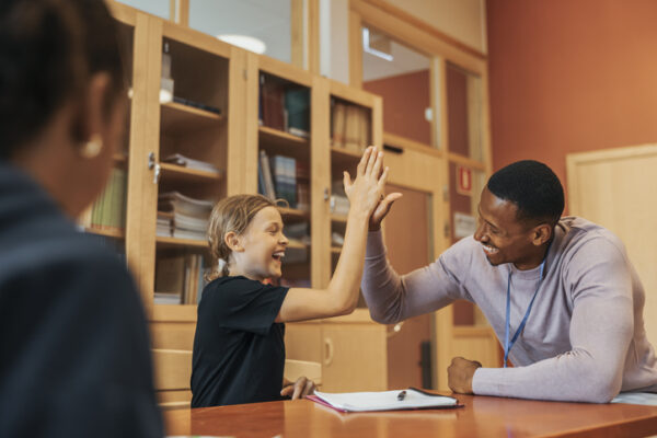 Happy boy giving high-five to male counselor sitting at table in middle school