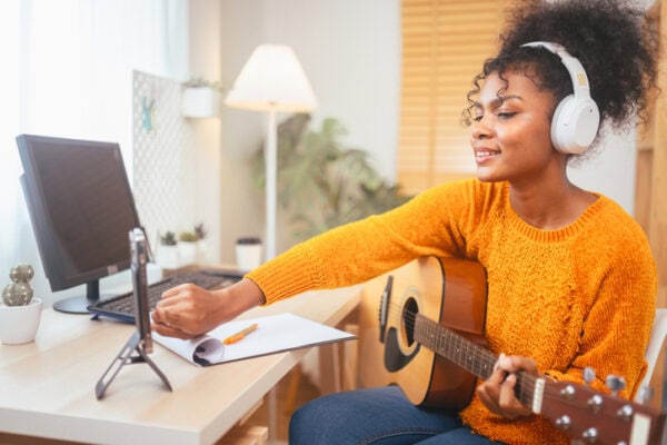 Woman playing guitar and taking notes at home studio. Online music lesson, creativity, and learning concept. Cozy workspace.