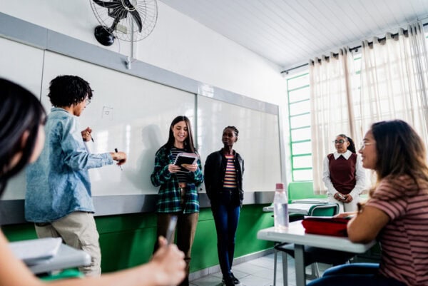 Students stand in front of the whiteboard, holding materials and presenting a school project to the class. Their classmates watch attentively from their desks. The scene highlights engagement, collaboration, and the development of communication skills in a classroom