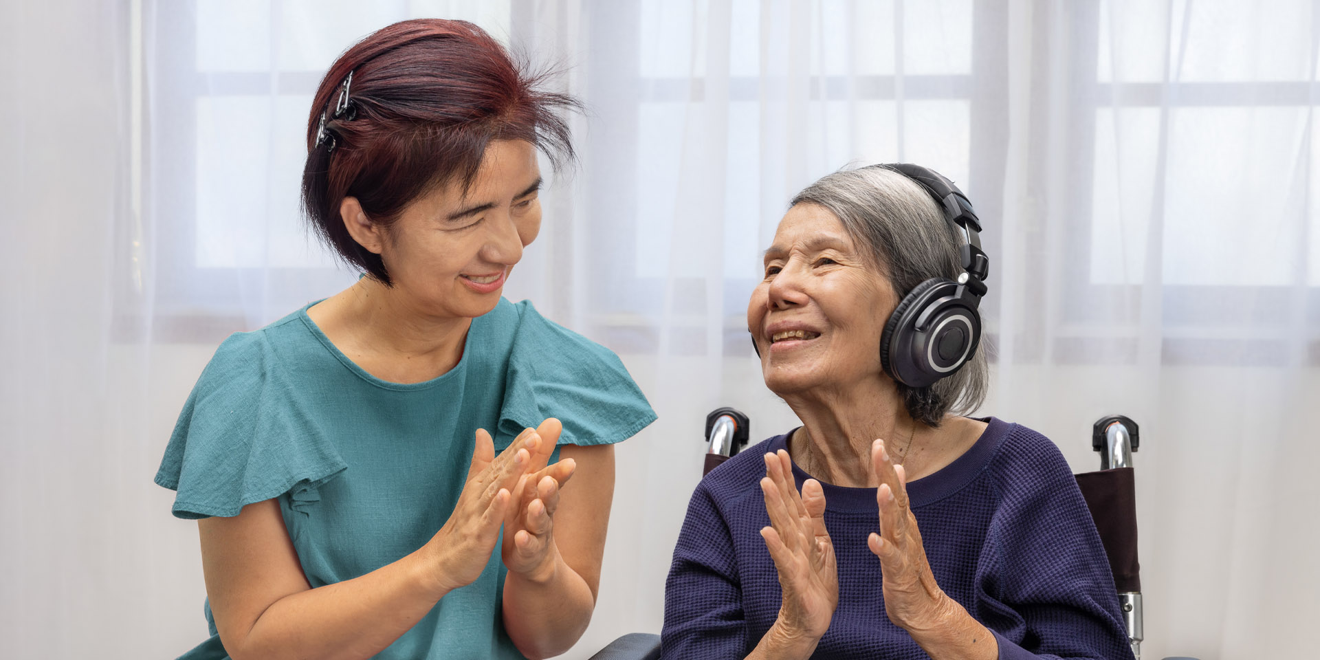 A middle-age woman smiling clapping with an elderly woman wearing headphones smiling and clapping